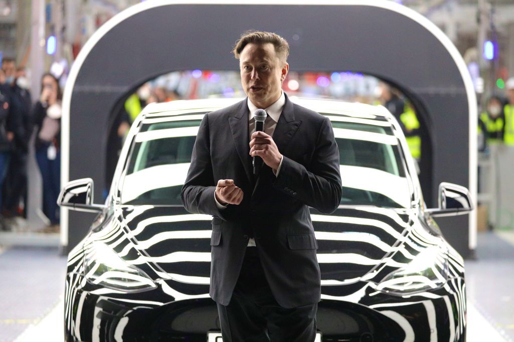 Elon Musk con traje negro, sosteniendo un micrófono, de pie frente a un auto Tesla en un evento corporativo.