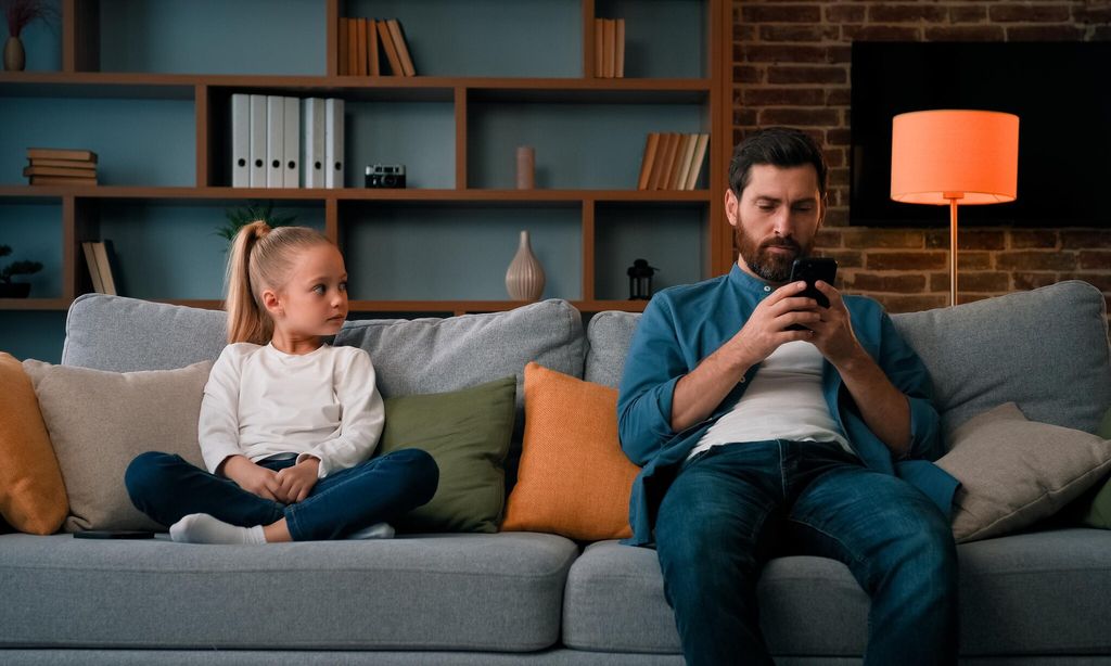 padre e hija sentados en el sof el padre mira el movil mientras la ni a lo observa aburrida