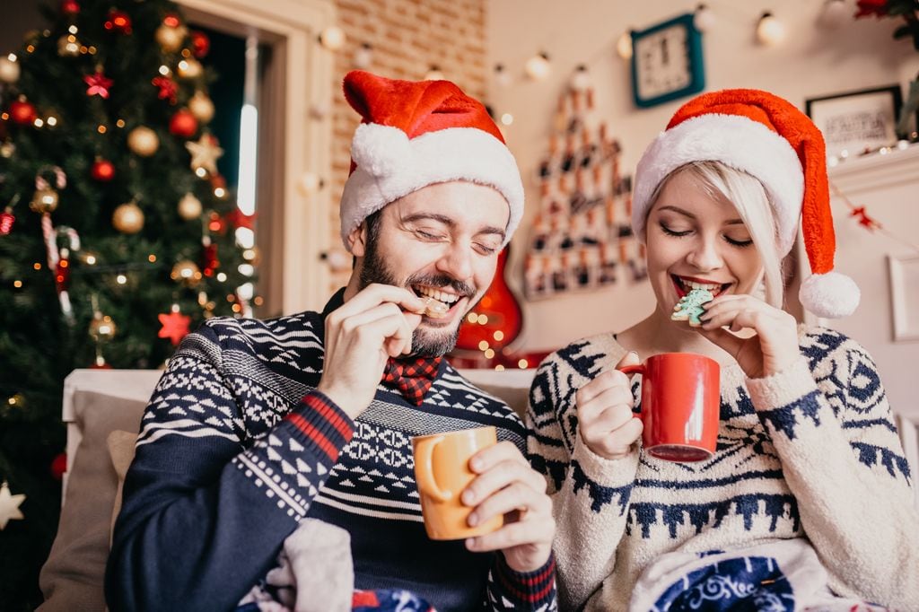 pareja con gorros de Papá Noél comiendo galletas y chocolate caliente en casa, en Navidad