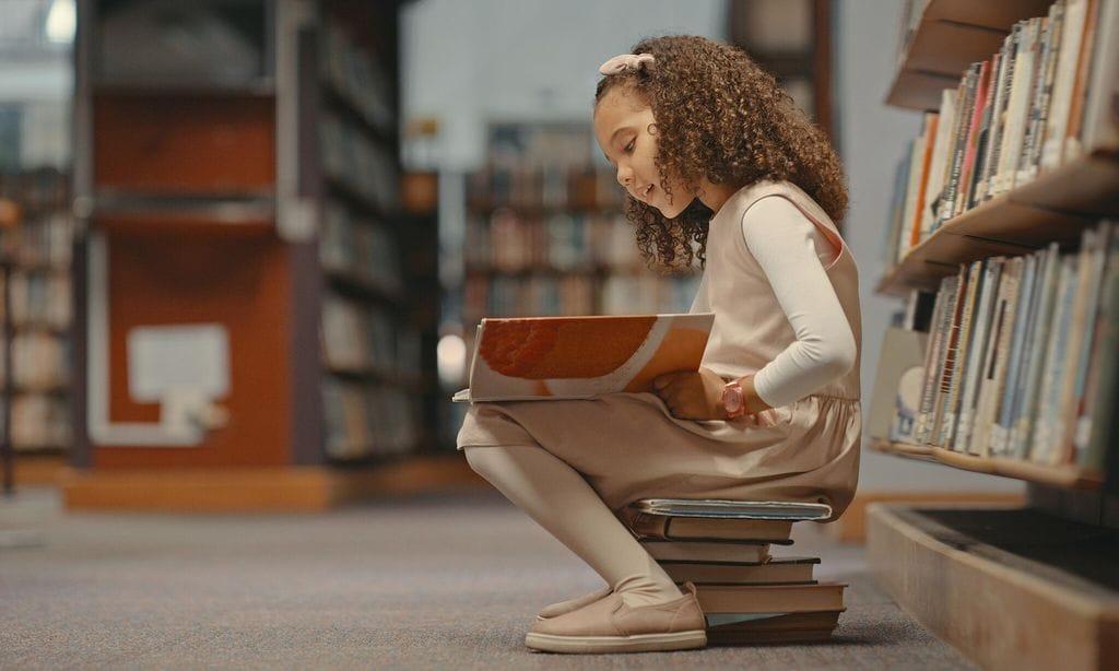 young girl sitting on books in the library and reading a book cute girl with curly hair doing her project female alone and doing research for a project