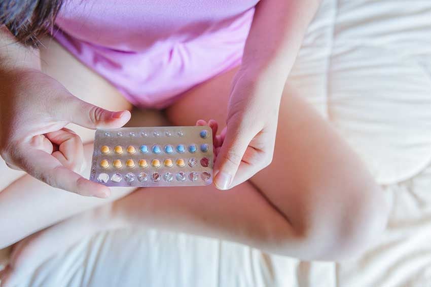 Mujer sentada en la cama con las pastillas anticonceptivas en la mano