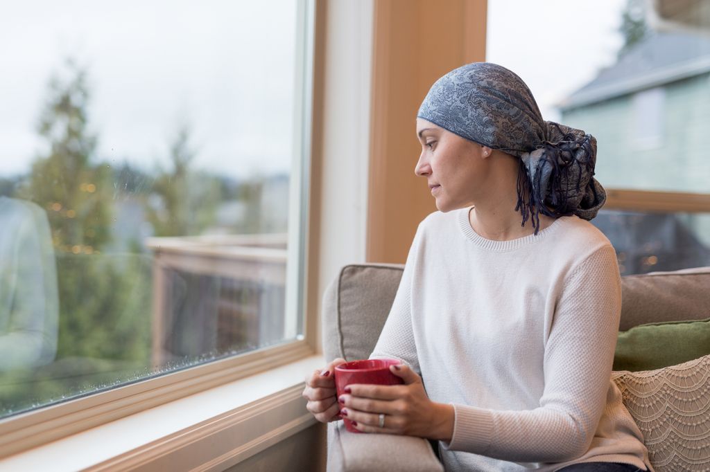 mujer con cáncer, con la cabeza cubierta con un pañuelo, tomando un café y mirando por la ventana