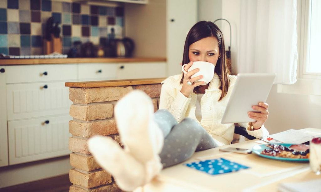 mujer sentada en la cocina de su hogar abrigada con taza de caf en mano y tablet