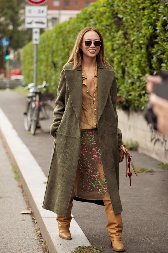 Niki Wu Jie con camisa y botas marrones, falda étnica y abrigo largo verde en la Milan Fashion Week 2024