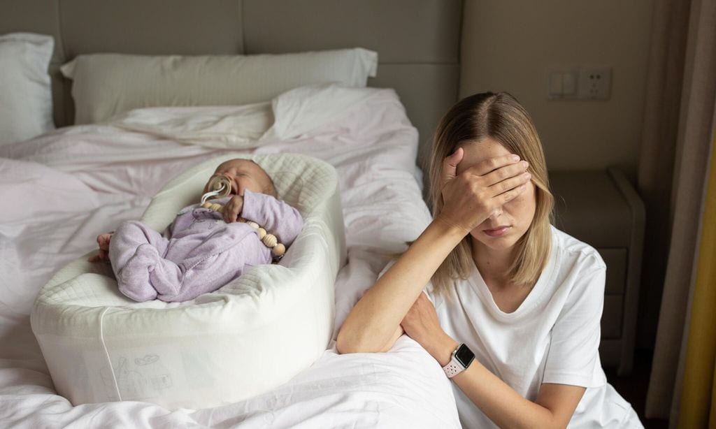 mujer con la mano tapando su cara apoyada en la cama con su beb al lado