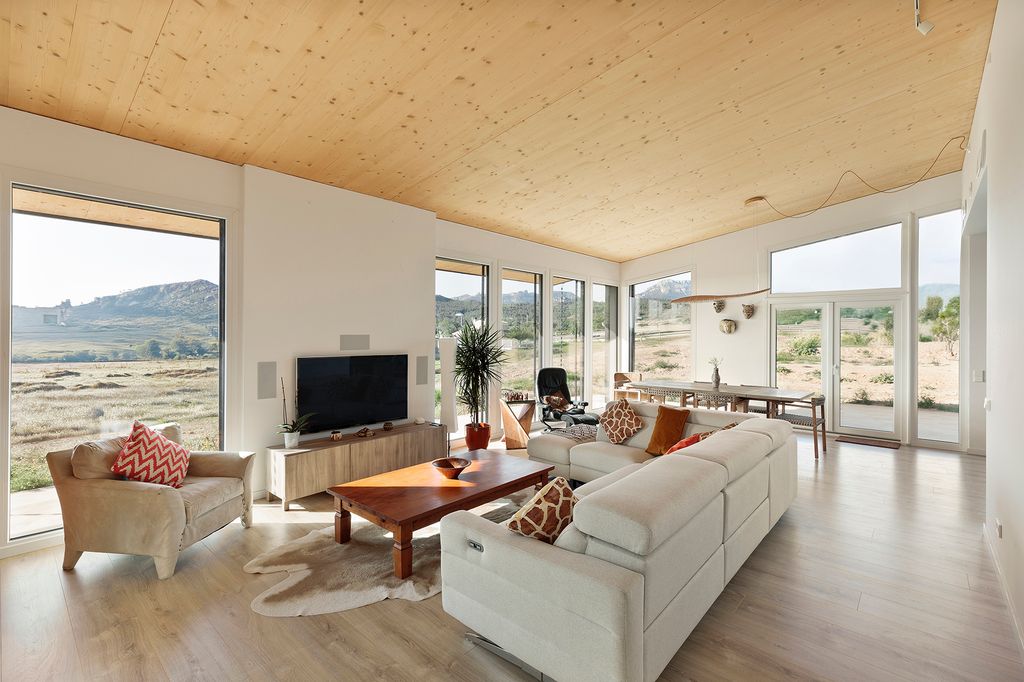 Salón comedor con grandes ventanales, sofá blanco, techo y suelo de madera, mueble televisor y mesa de centro de madera