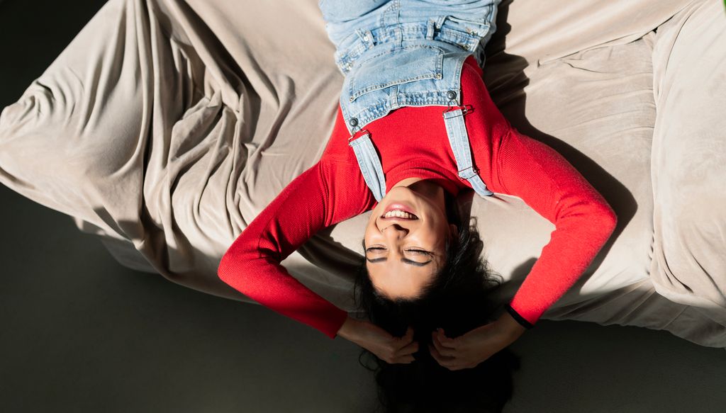 Mujer joven tumbada sobre un sofá sonriendo y feliz, con un jersey rojo y un peto vaquero, dándole la luz en la cara