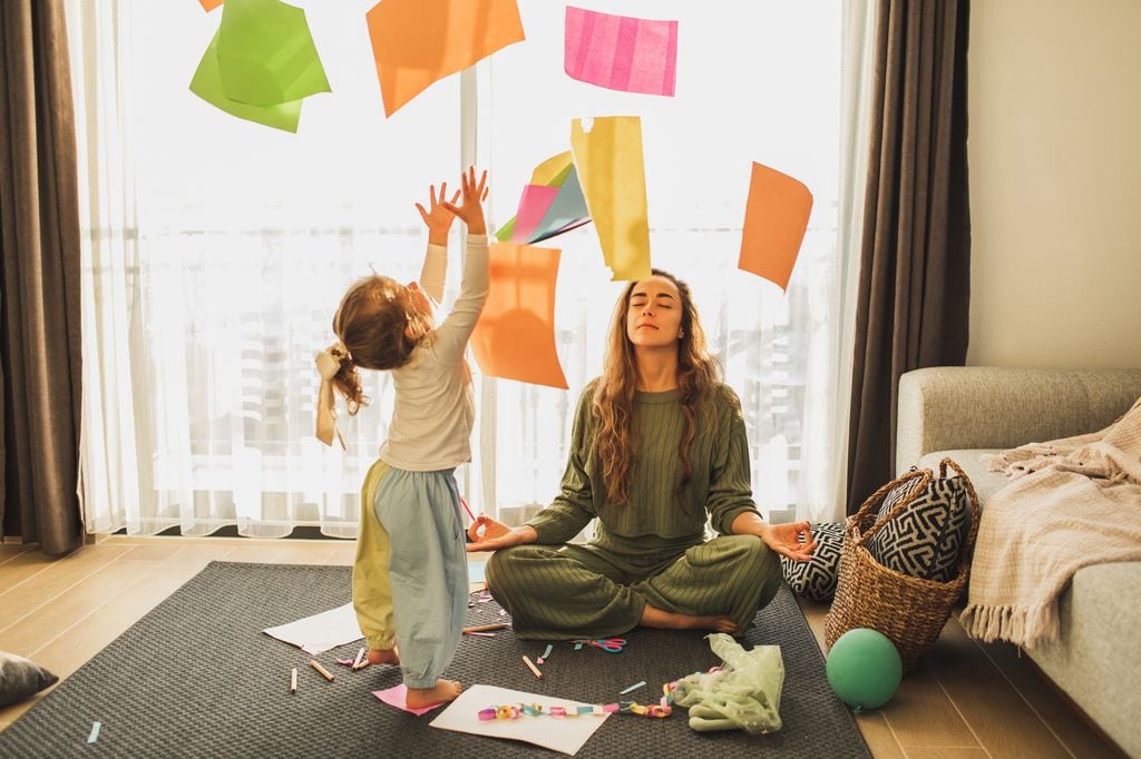 mujer meditando en casa mientras su hija juega lanzando unos papeles al aire