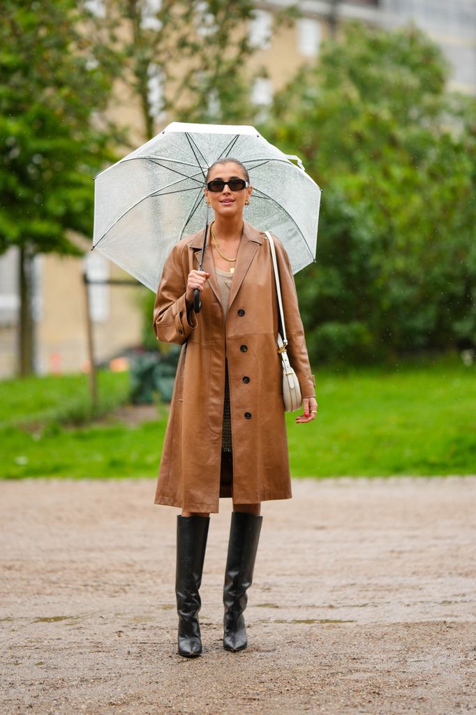 Una invitada lleva un paraguas blanco transparente, gafas de sol negras, un abrigo largo de cuero camel brillante, collares de oro, un vestido corto de lana trenzada beige de Prada, botas / botas altas de tacón de punta de cuero negro brillante, un bolso de hombro de cuero blanco mate latte, anillos de oro , fuera de OperaSport, durante la Semana de la Moda de Copenhague Primavera/Verano 2024 el 09 de agosto de 2023 en Copenhague, Dinamarca.