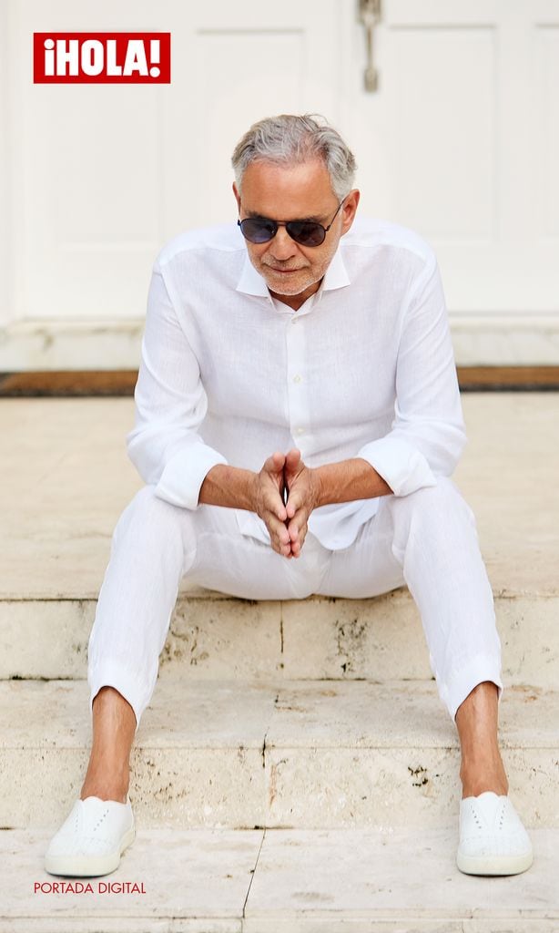 Andrea Bocelli, vestido completamente de blanco y con gafas de sol, está sentado en las escaleras de su casa en Miami, reflejando serenidad y elegancia.