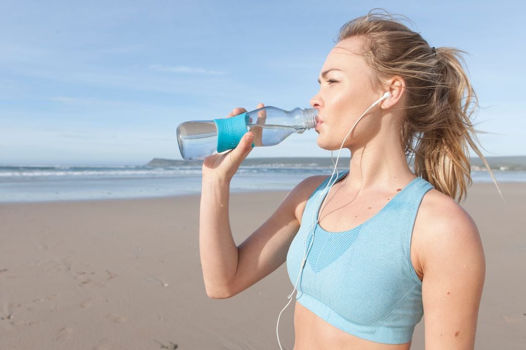 mujer en la playa bebiendo agua de una botella, con unos auriculares