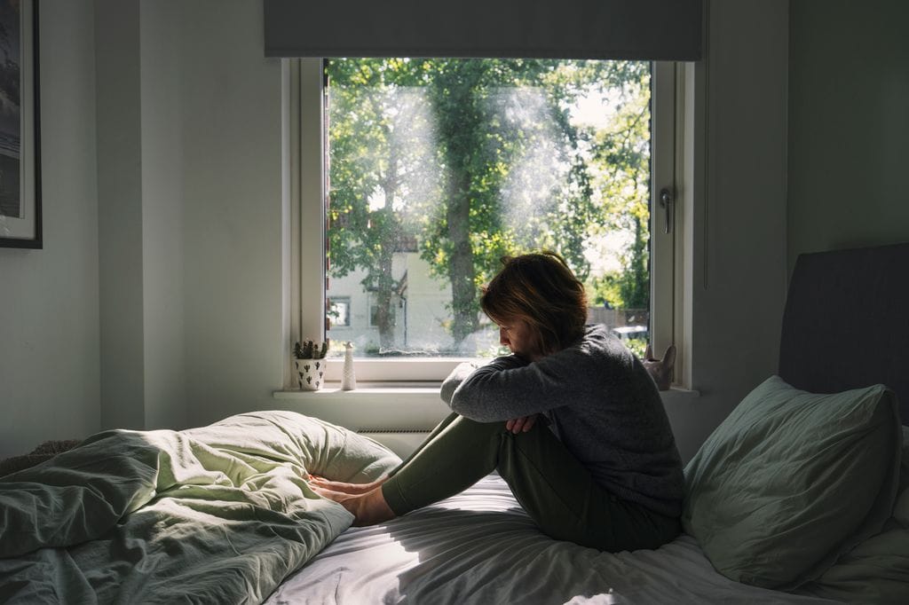 mujer pensativa sentada en la cama, junto a la ventana