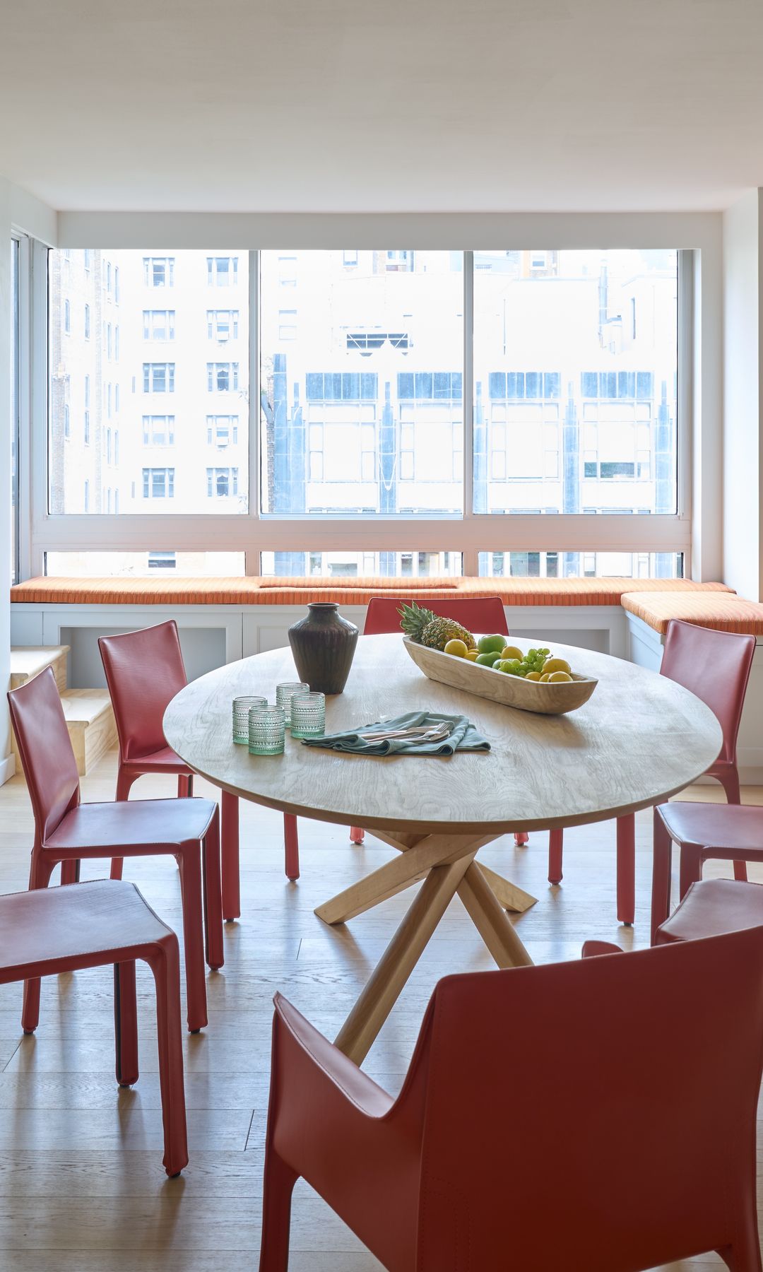 Comedor con mesa de madera, sillas rojas y banco con cojín naranja junto a la ventana