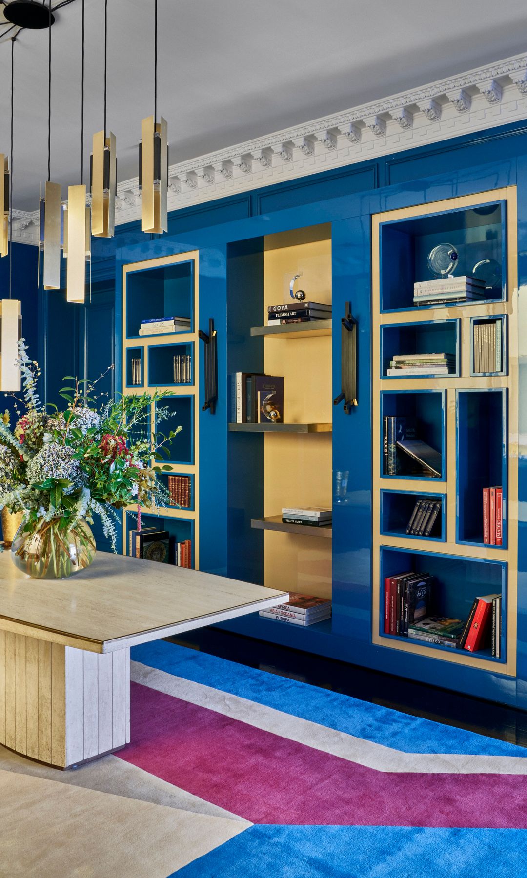 Mueble a medida azul con trasera amarilla y molduras decorativas en el techo, mesa de comedor de mármol , jarrón con flores y lámpara de suspensión con pantallas doradas