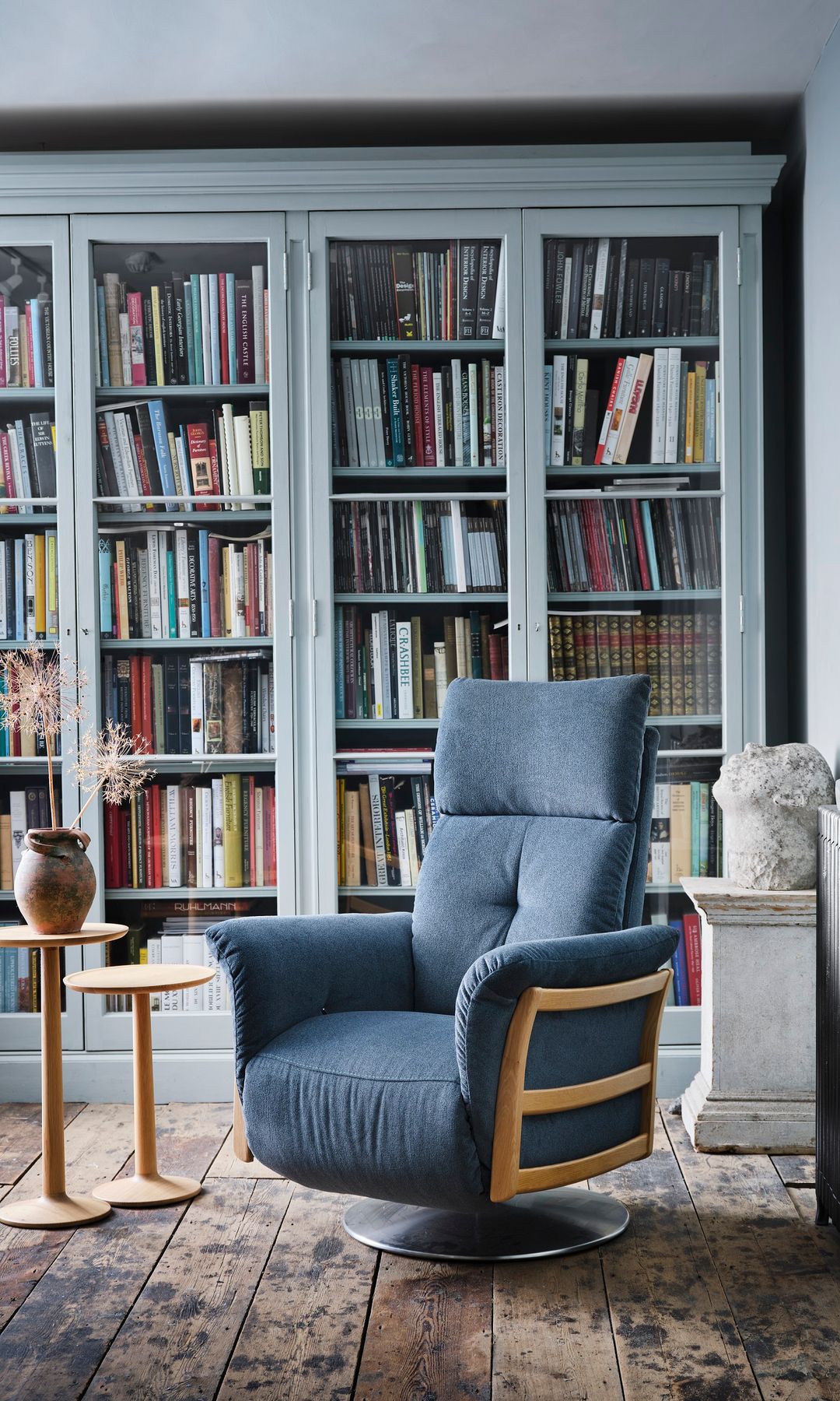 Rincón de lectura con butaca gris, suelo de madera, mesas auxiliares y librería en color gris con puertas de cristal llena de libros