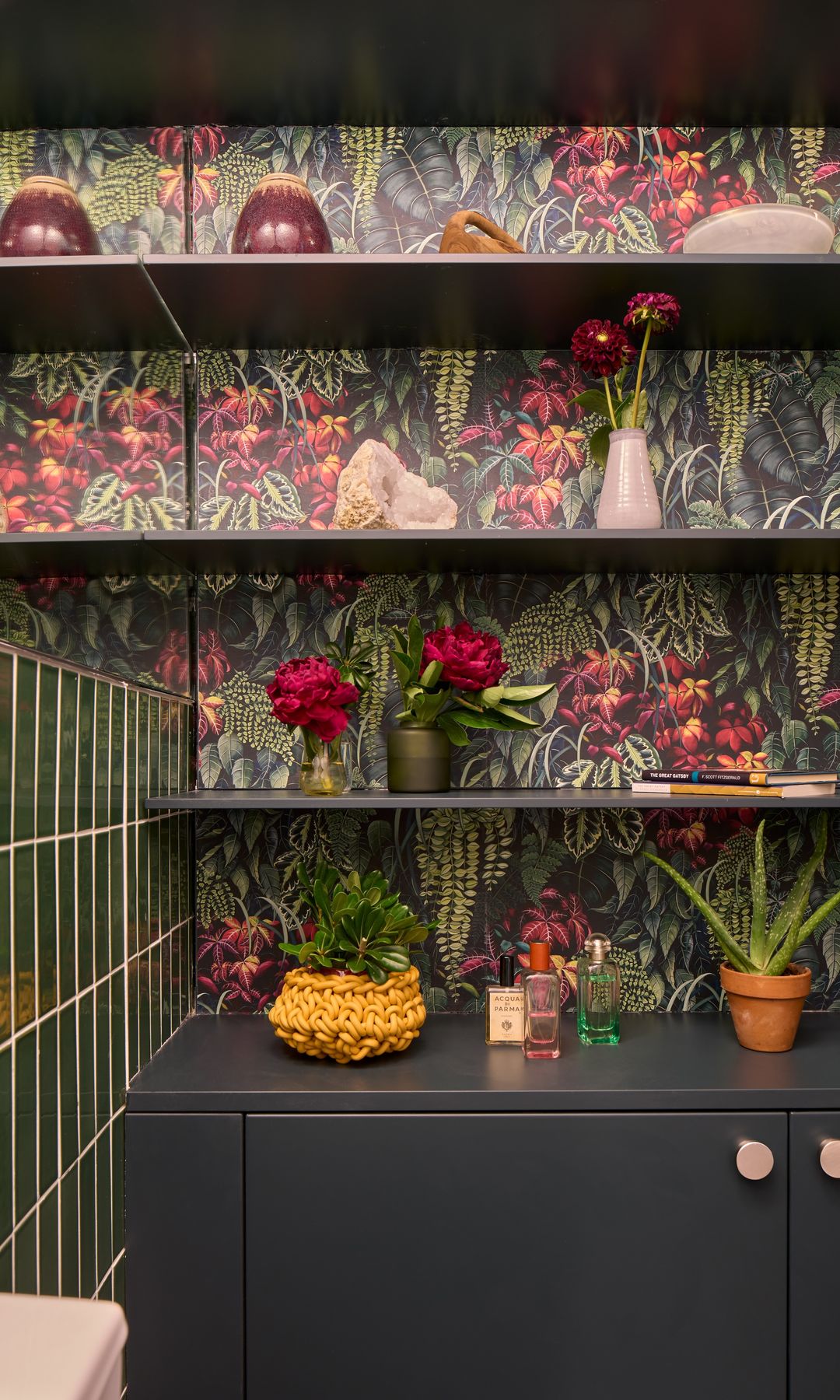 Baño con papel pintado con motivos vegetales, azulejos verdes rectangulares y mueble verde