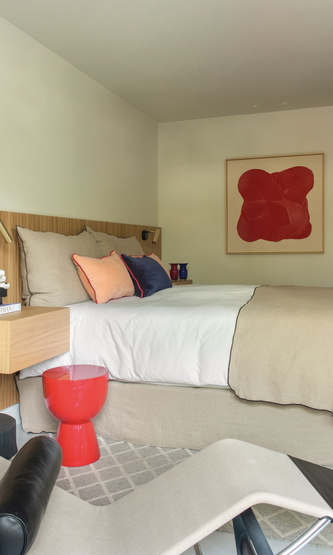 Dormitorio con cabecero de listones de madera, ropa de cama y paredes crema, taburete rojo y cuadro rojo y crema