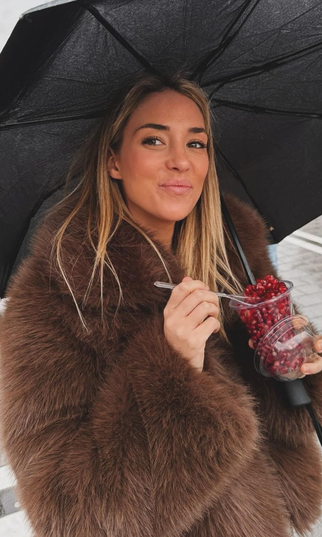 Alice Campello con abrigo de pelo marrón y paraguas comiendo fruta en Estambul