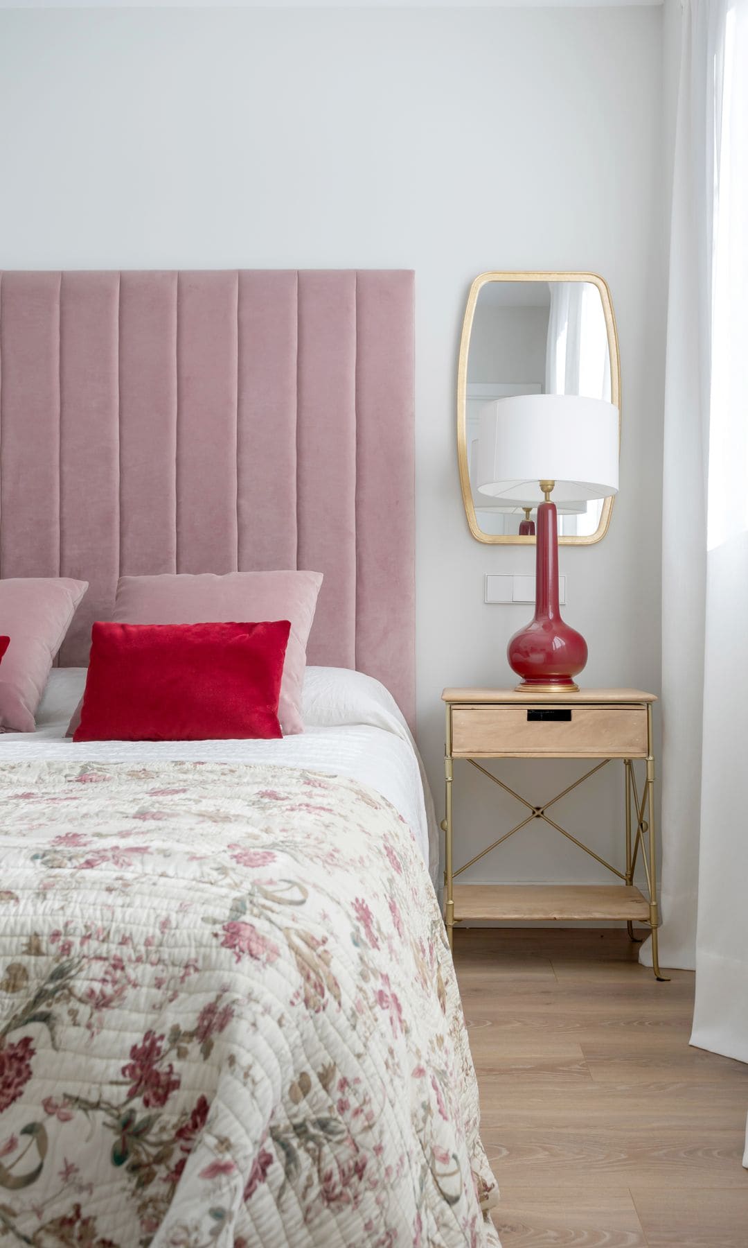 Dormitorio con cabecero tapizado rosa, colcha con motivos florales, mesita de noche de madera, lámpara de sobremesa roja y blanca y cojines rosas y rojos, 