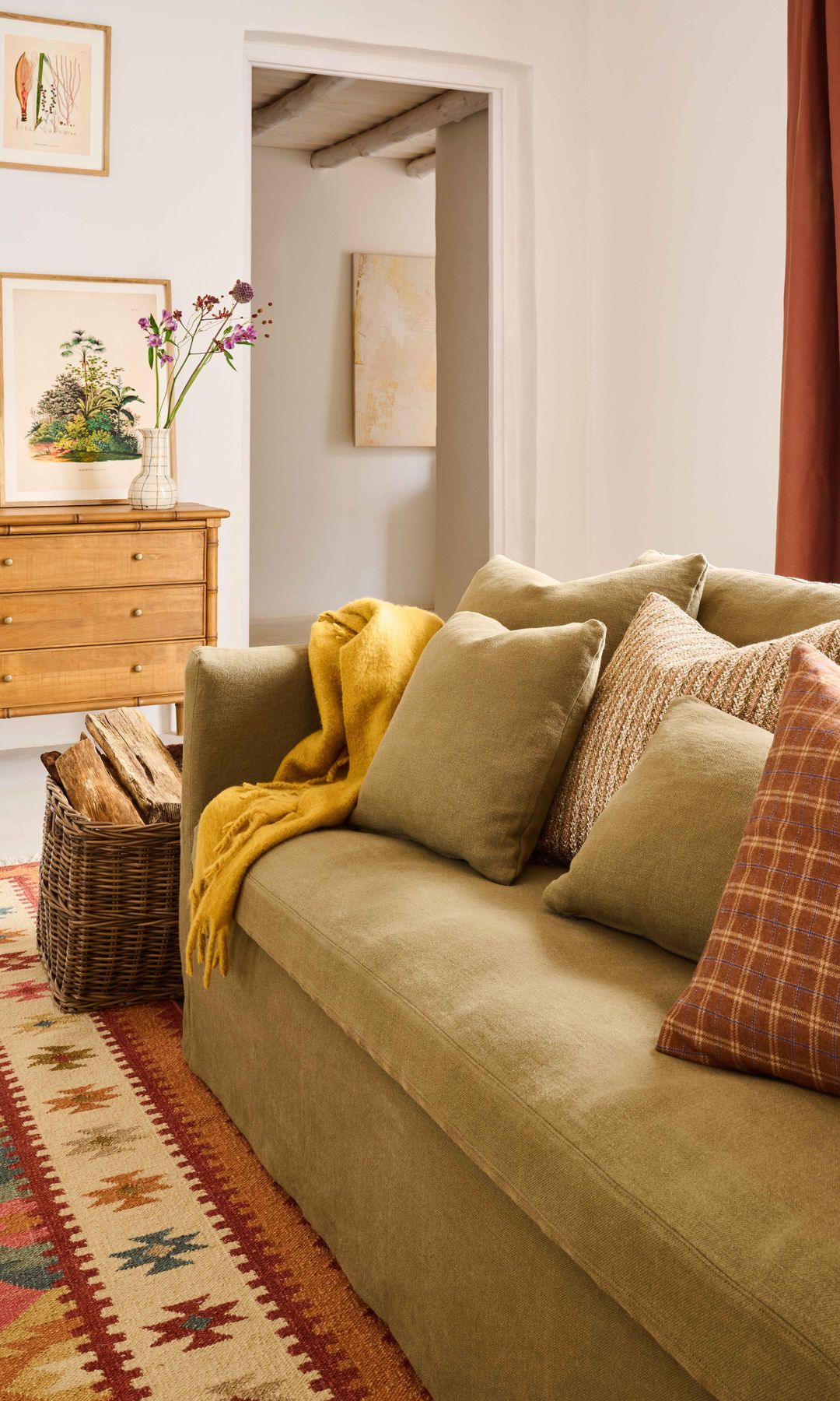 Salón con sofá beige lleno de cojines, cesto con leña, alfombra, cómoda de madera, cuadros y jarrón con flores