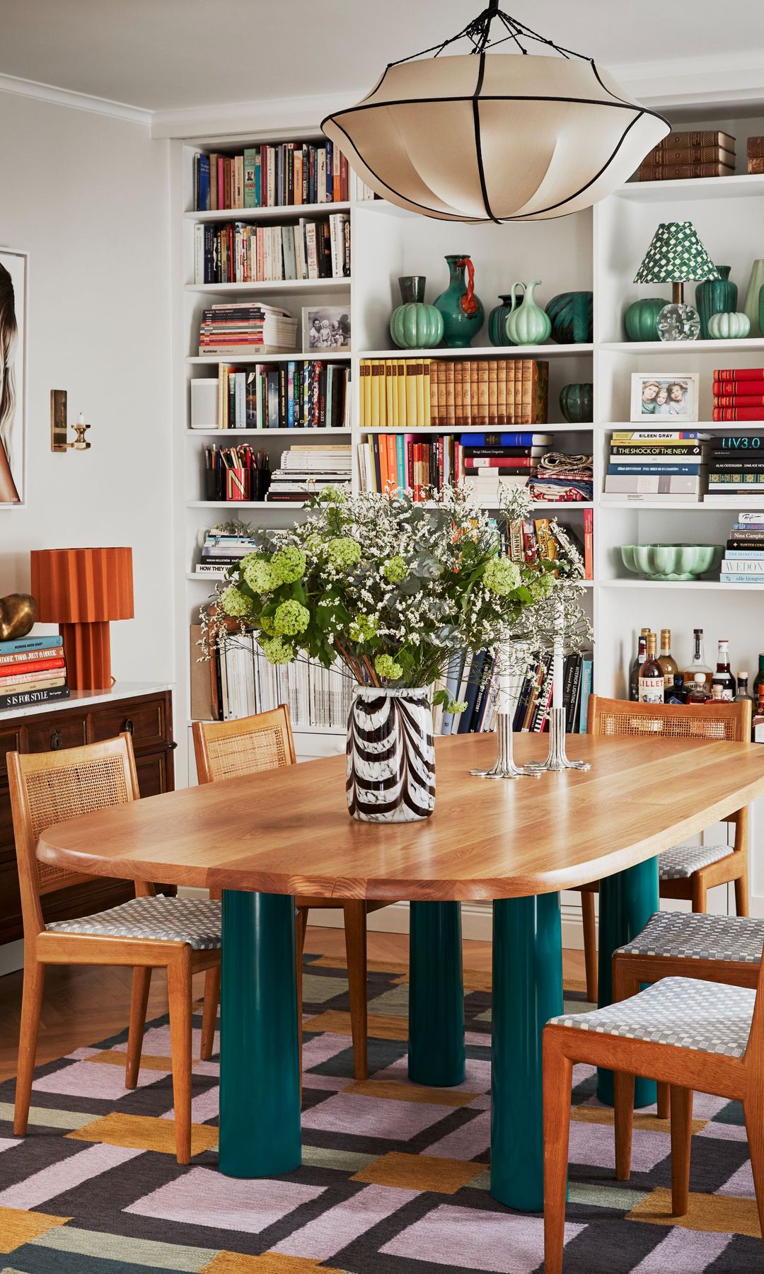 Comedor con mesa de madera con patas verdes, arreglo floral, alfombra geométrica de los setenta y aparador 