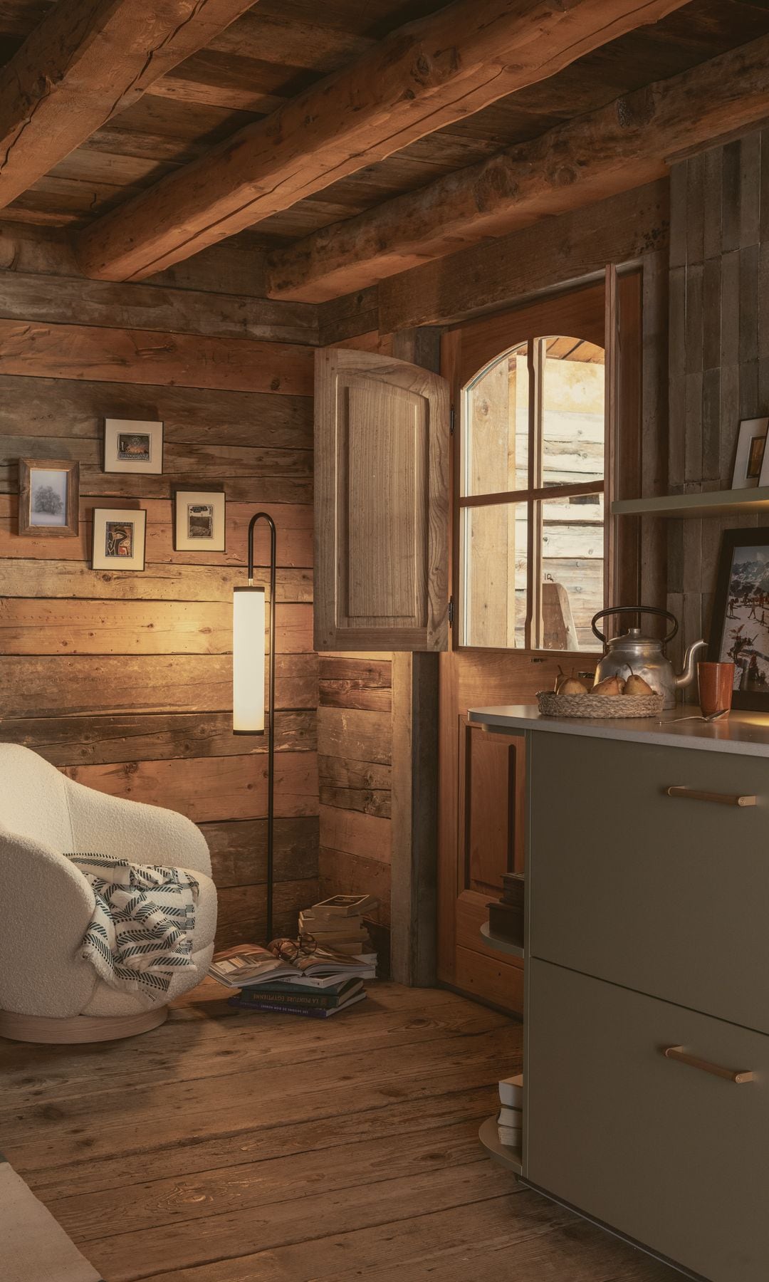 Cabaña con sillón blanco, suelo de madera, vigas de madera, mueble verde, tetera, cuadros pequeños en la pared