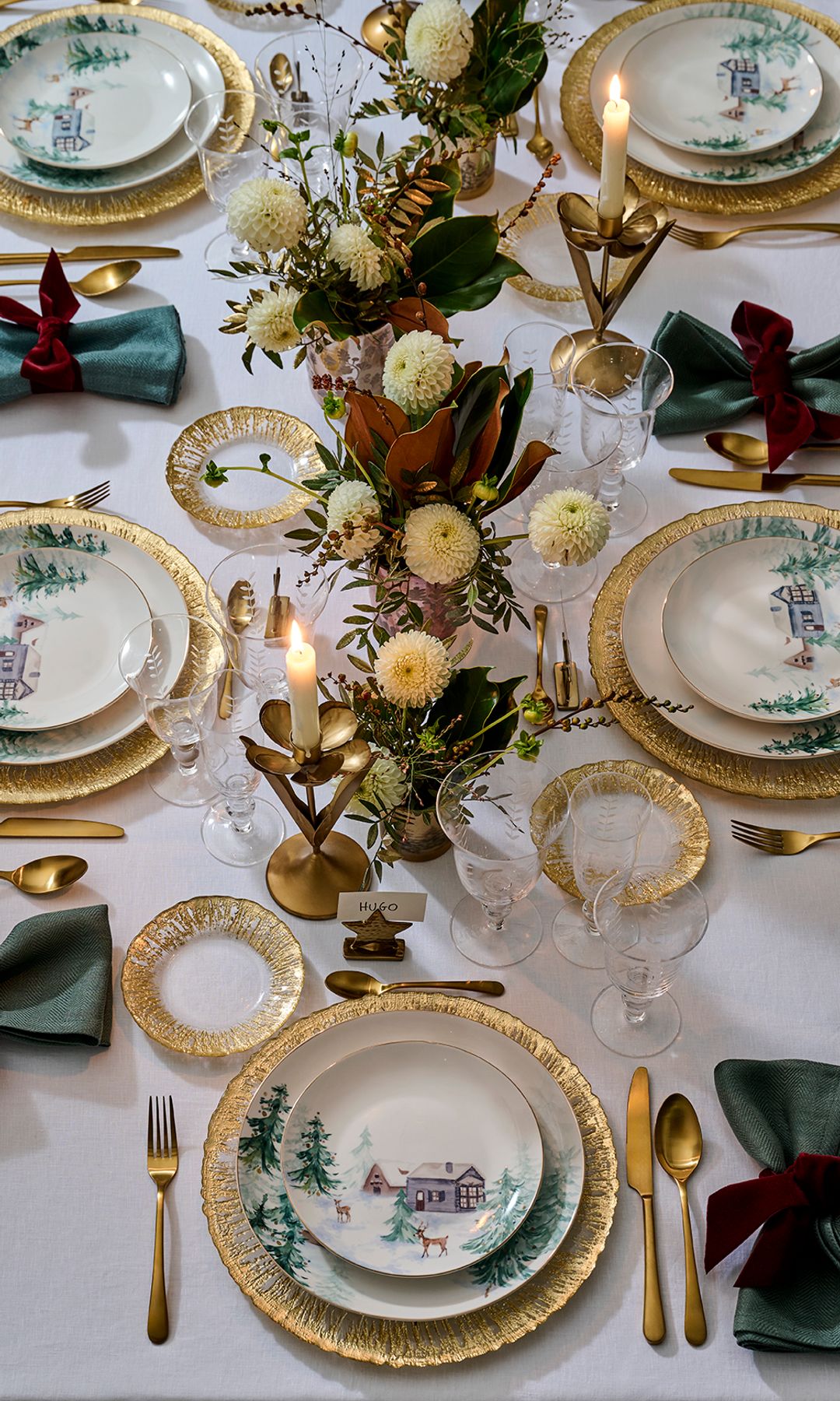 Mesa Navidad con bajoplatos y cubiertos dorados, mantel blanco, servilletas verdes y vajilla con motivos navideños