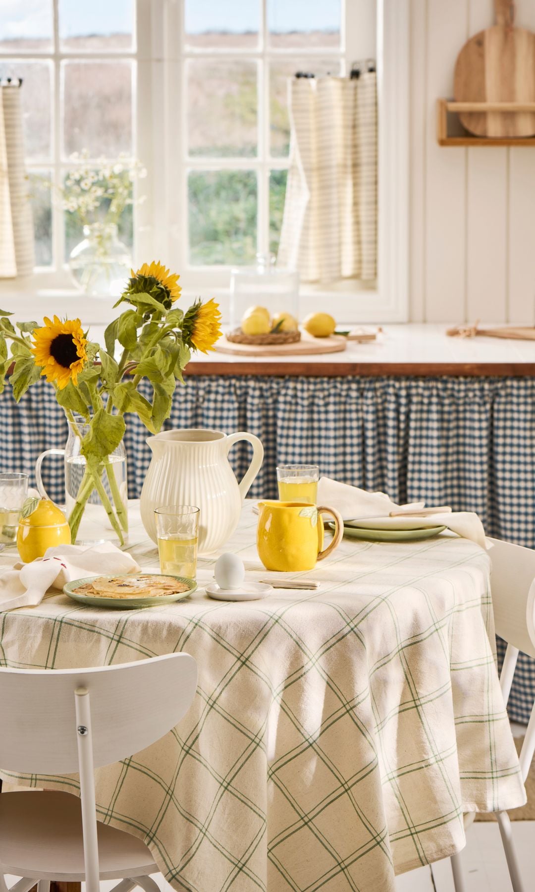 Cocina con mesa de comedor redonda con mantel, tazas amarillas, jarra, jarrón con tulipanes, cortinillas de cuadros tapando el fregadero