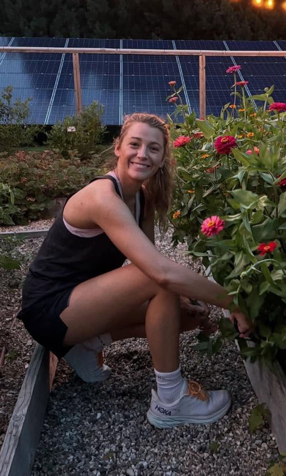 Blacke Lively con shorts y camiseta de tirantes negra, zapatillas Hoka y coleta en un jardín cortando flores