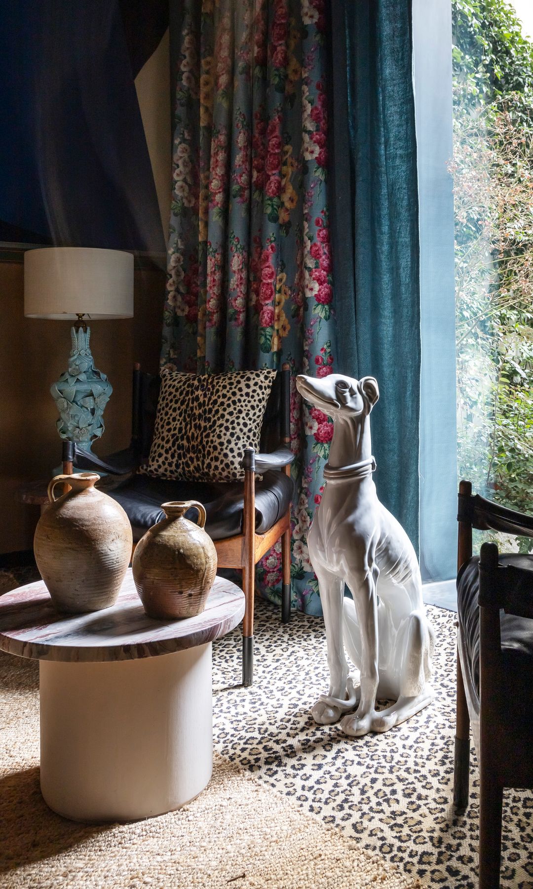 Rincón del salón con alfombra de animal print, cortinas de flores, perro de porcelana grande, cojín de leopardo, mesa auxiliar redonda y dos jarrones de barro