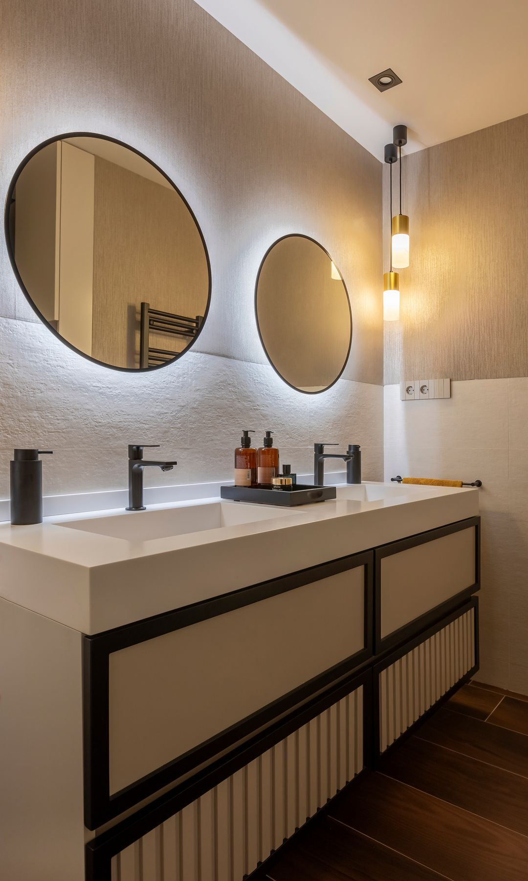 Baño con espejos redondos retroiluminados, mueble bajolavabo beige con detalles negros