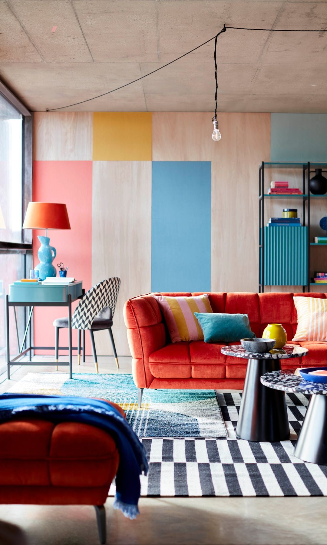 Salón con paredes de colores, sofá rojo, alfombra de rayas negras y blancas y zona de despacho con escritorio azul, lámpara de sobremesa con el pie azul y la pantalla roja y silla con estampado de damero