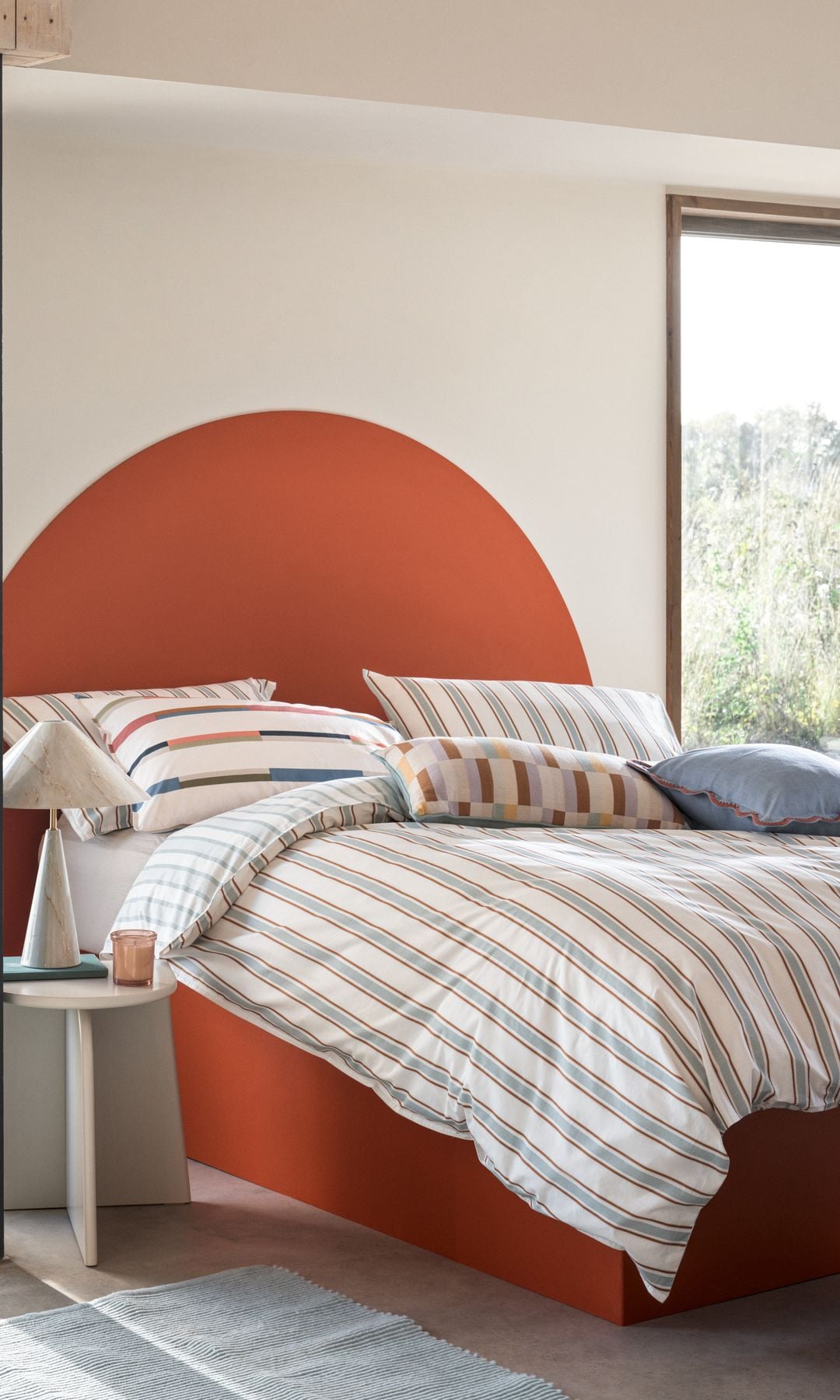 Cama tapizada naranja con ropa de cama a rayas y puff morado 