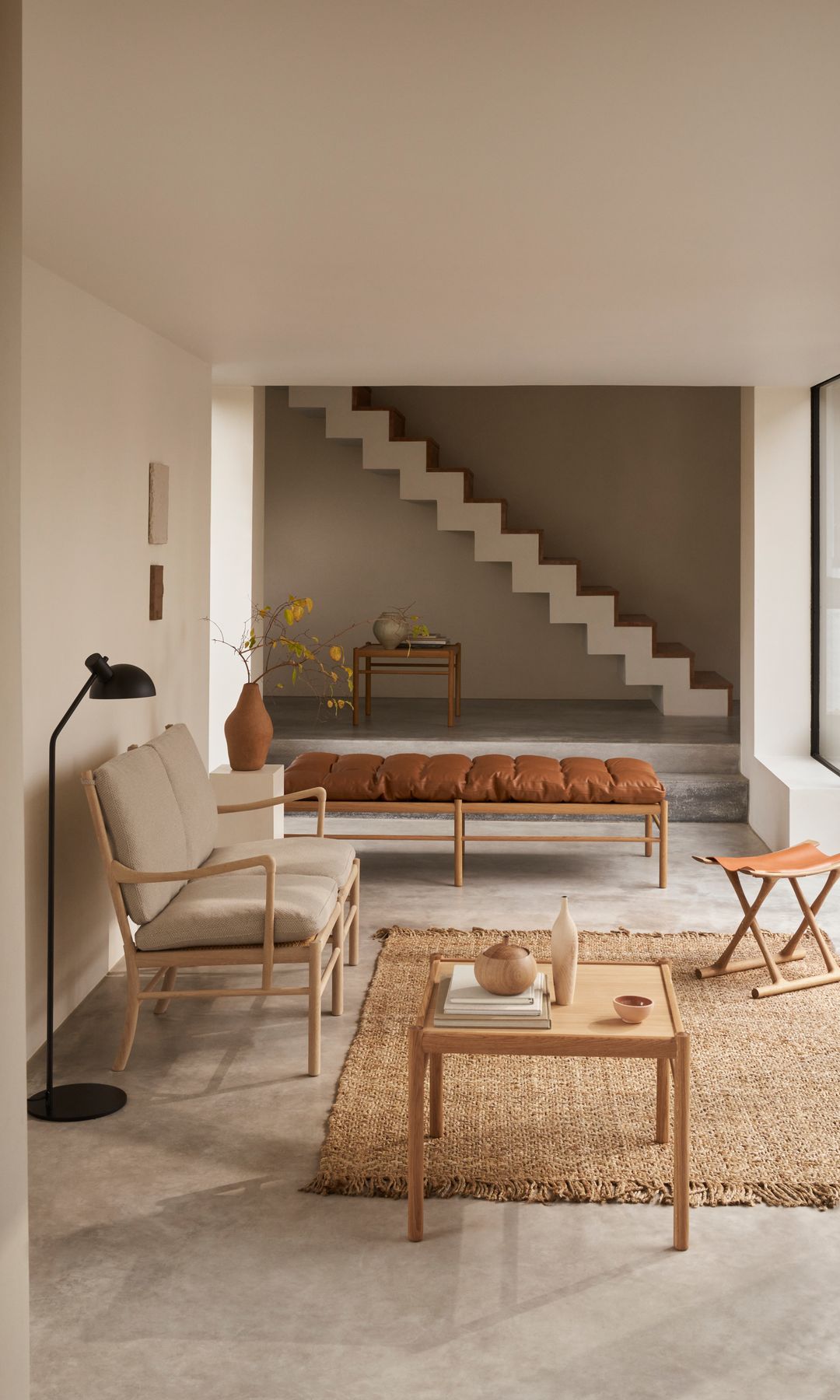 Salón con escalera al fondo, alfombra de fibras vegetales, sofá beige, lámpara de pie negra y gran ventanal con carpintería negra con salida al jardín 