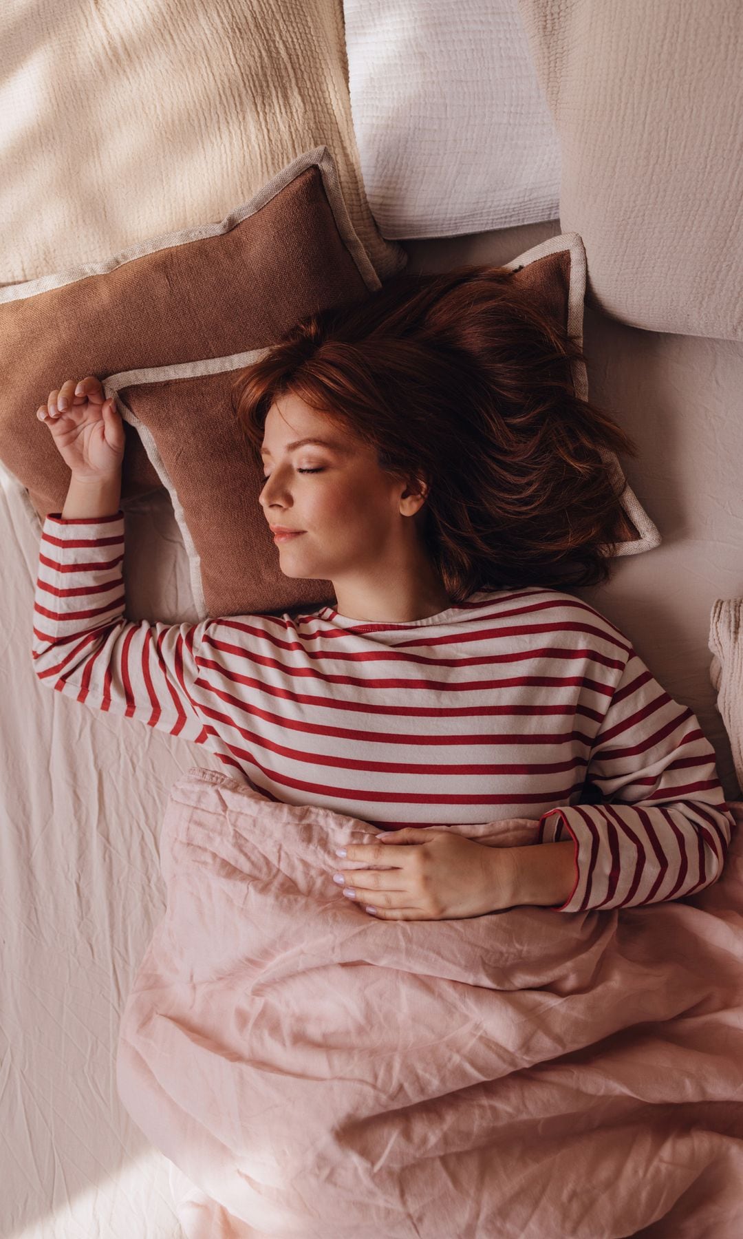 Mujer joven durmiendo en una cama con una camiseta de rayas rojas, sonriendo, sobre unas almohadas