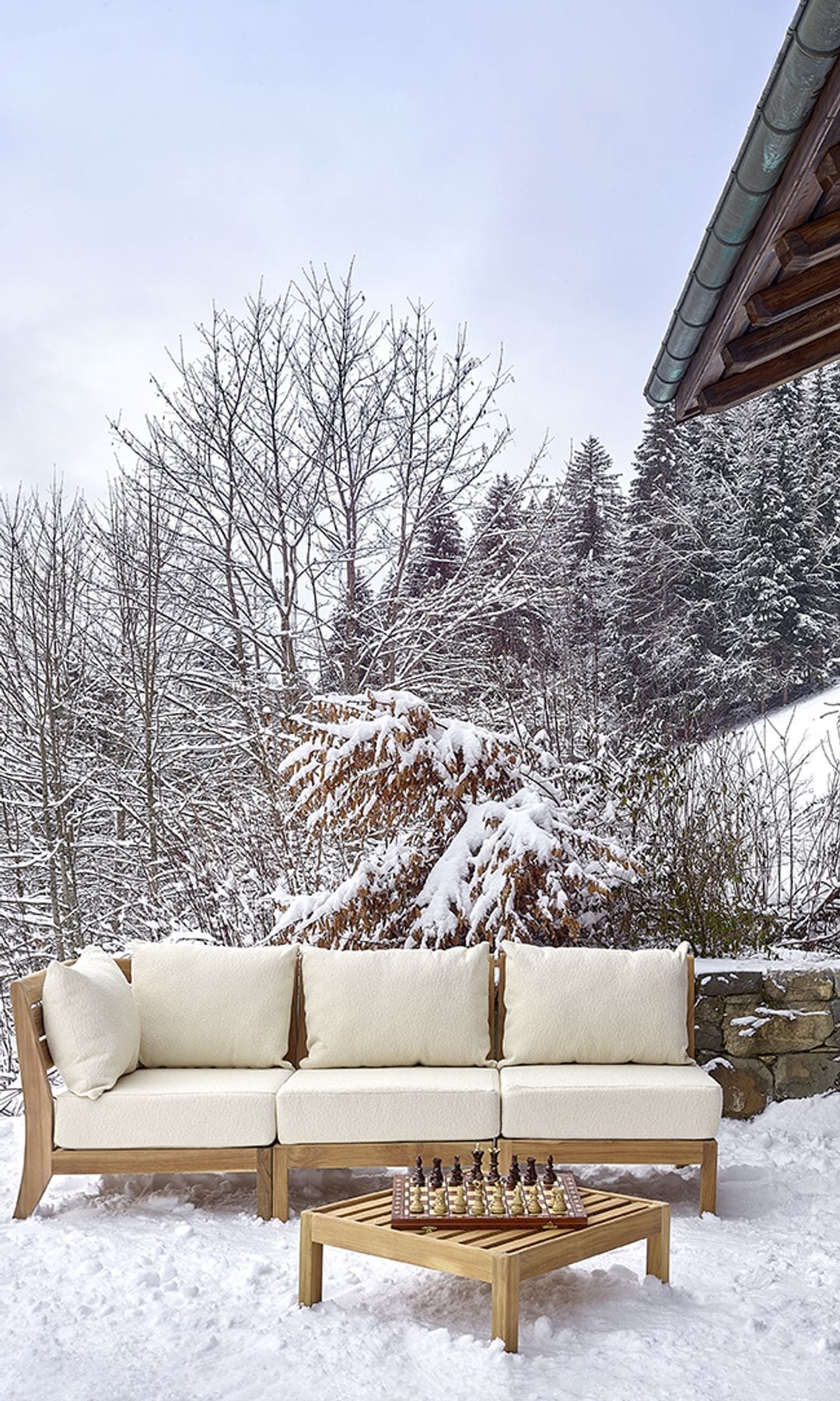 Cabaña nevada con abetos, sofá de madera con cojines blancos, mesa de centro de madera y ajedrez