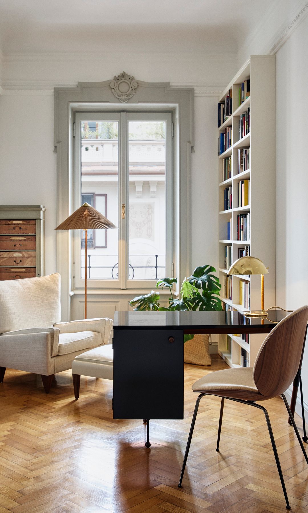 Despacho con suelo de madera, ventanas grandes con molduras, mesa negra, silla de madera, butaca blanca y lámpara de pie