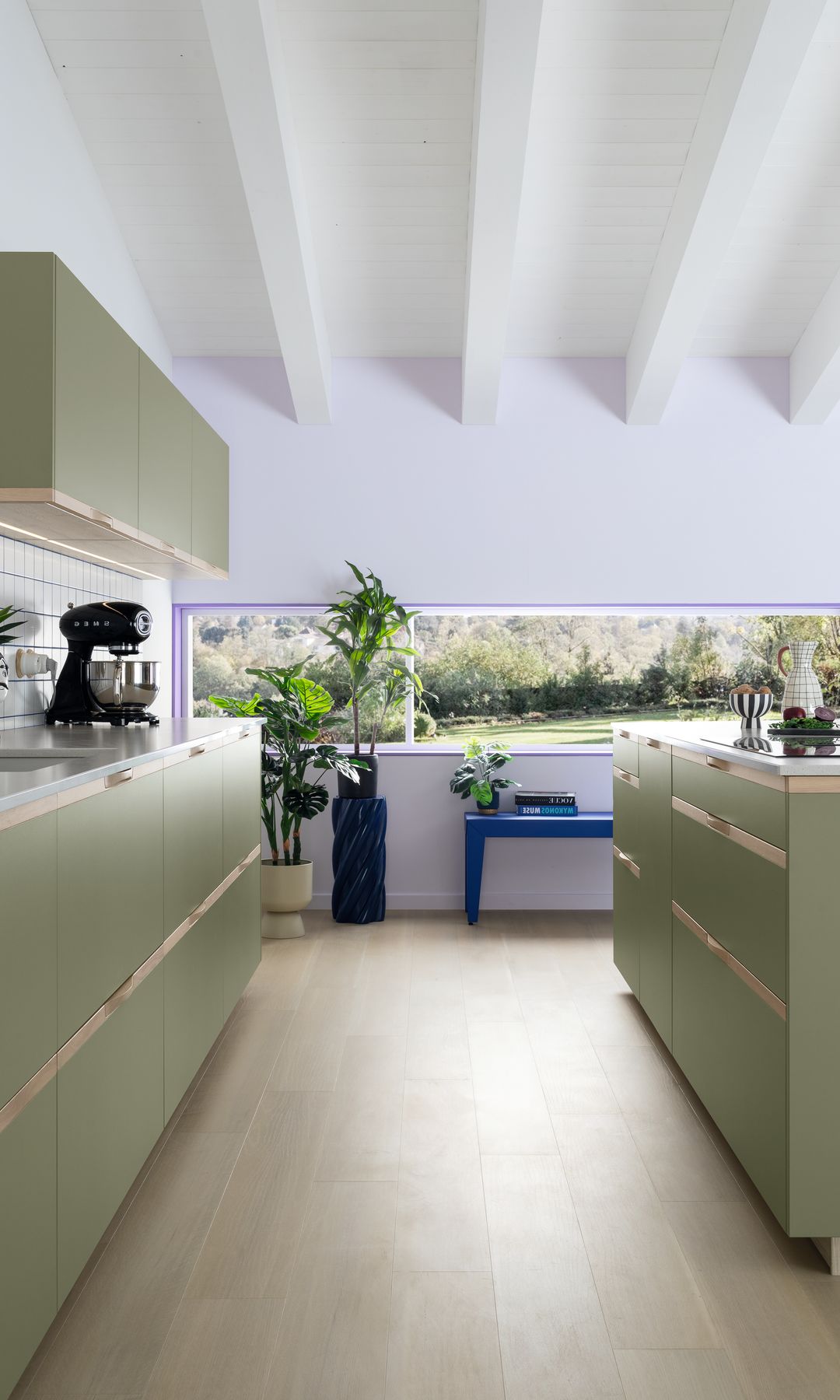 Cocina con muebles verde claro, suelo de madera, banco azul, plantas, batidora negra y vigas blancas en el techo 