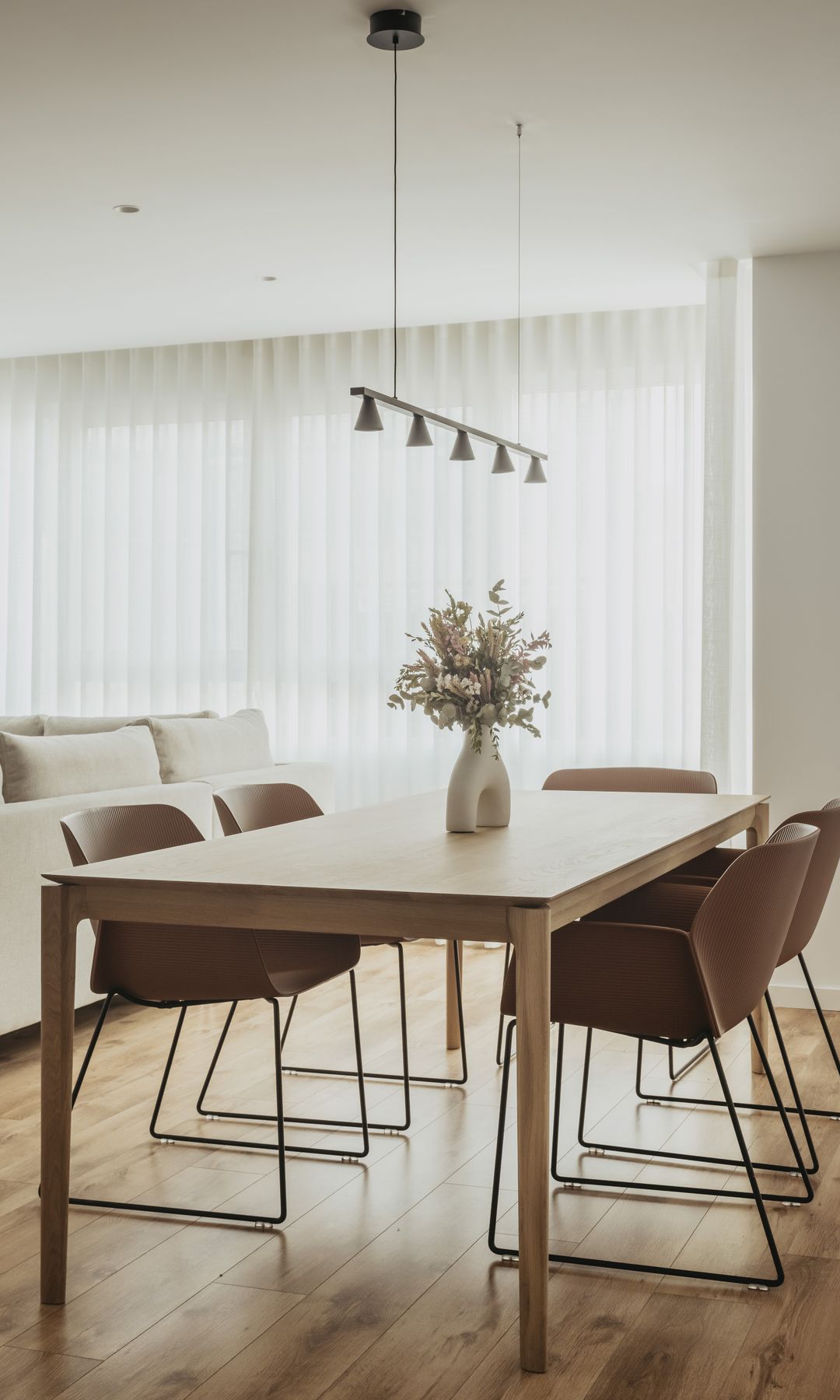 Salón comedor con suelo de madera, mesa de comedor rectangular de madera, sillas con brazos, lámpara de suspensión y jarrón escultura