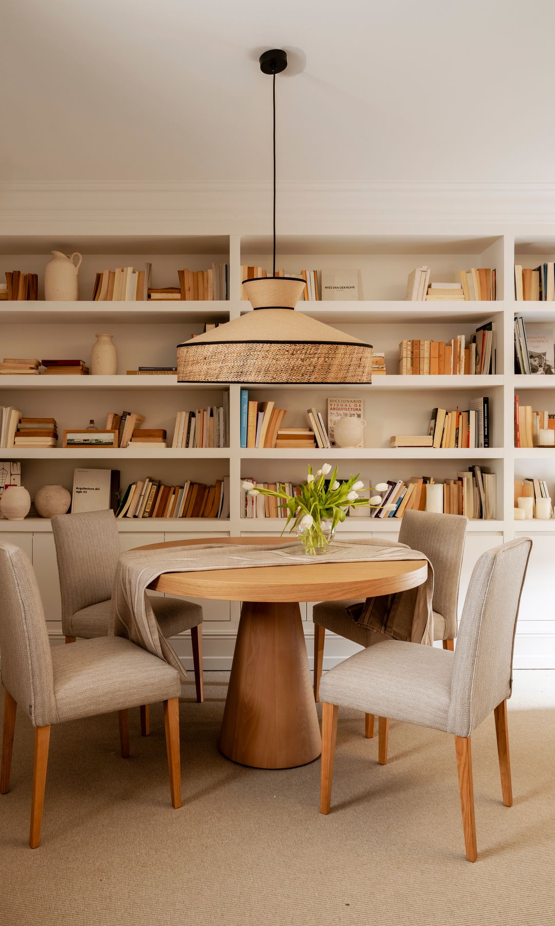 Comedor con mesa redonda de madera, sillas tapizadas, lámpara de techo y librería blanca con libros y adornos 