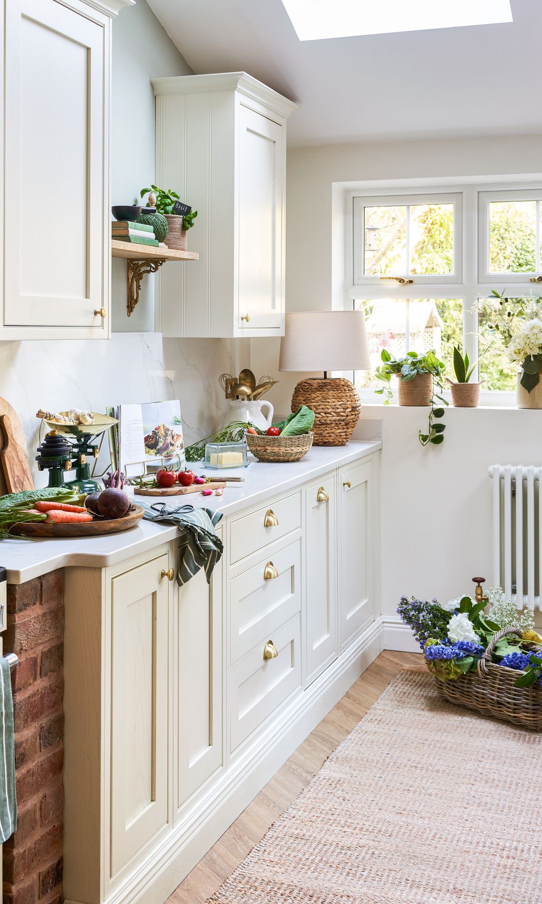 Cocina blanca con tiradores de concha, alfombra de fibras vegetales, plantas, lámpara de sobremesa, cestos y tablas