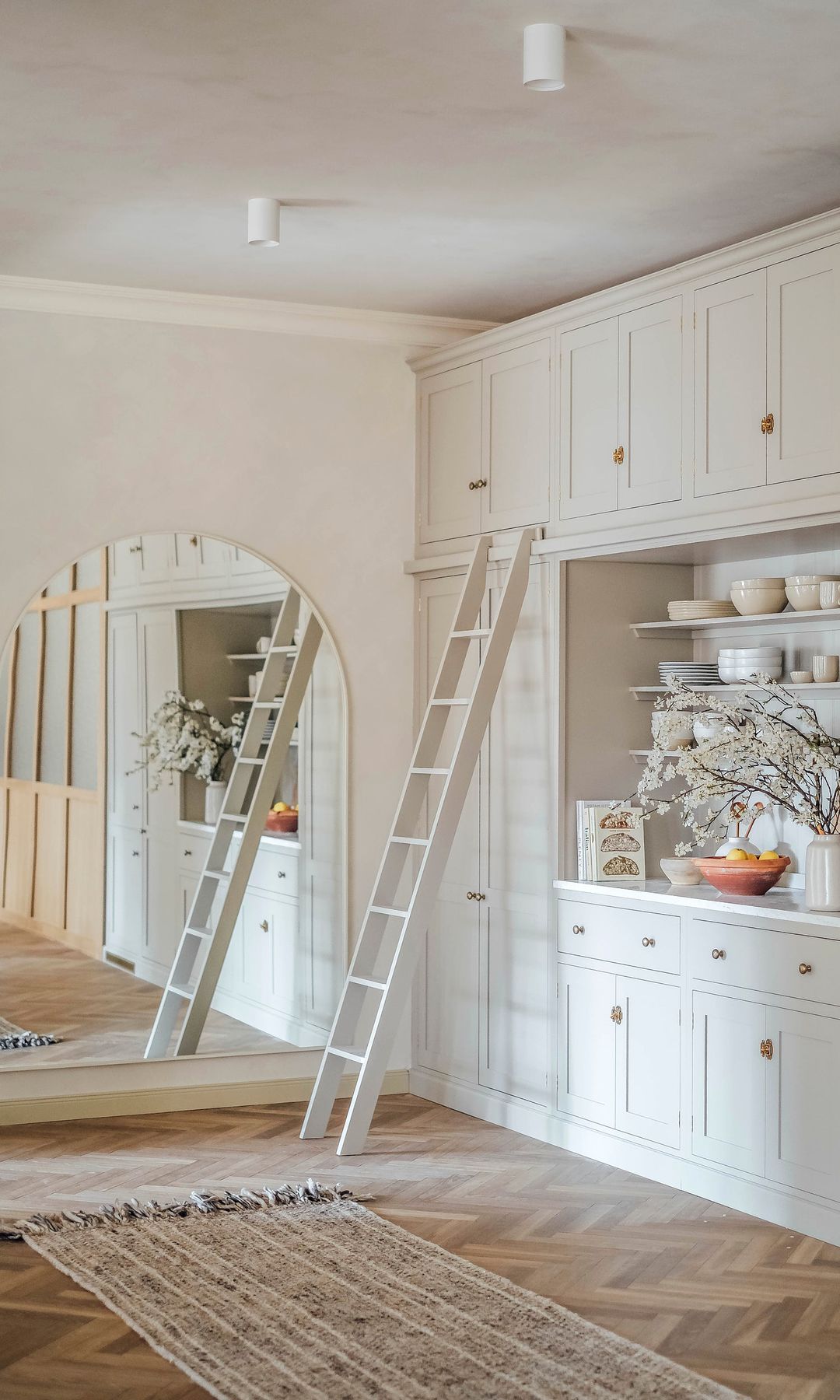 Cocina blanca con estantería abierta con escalera, espejo en forma de arco, alfombra de fibras vegetales y suelo de madera en espiga