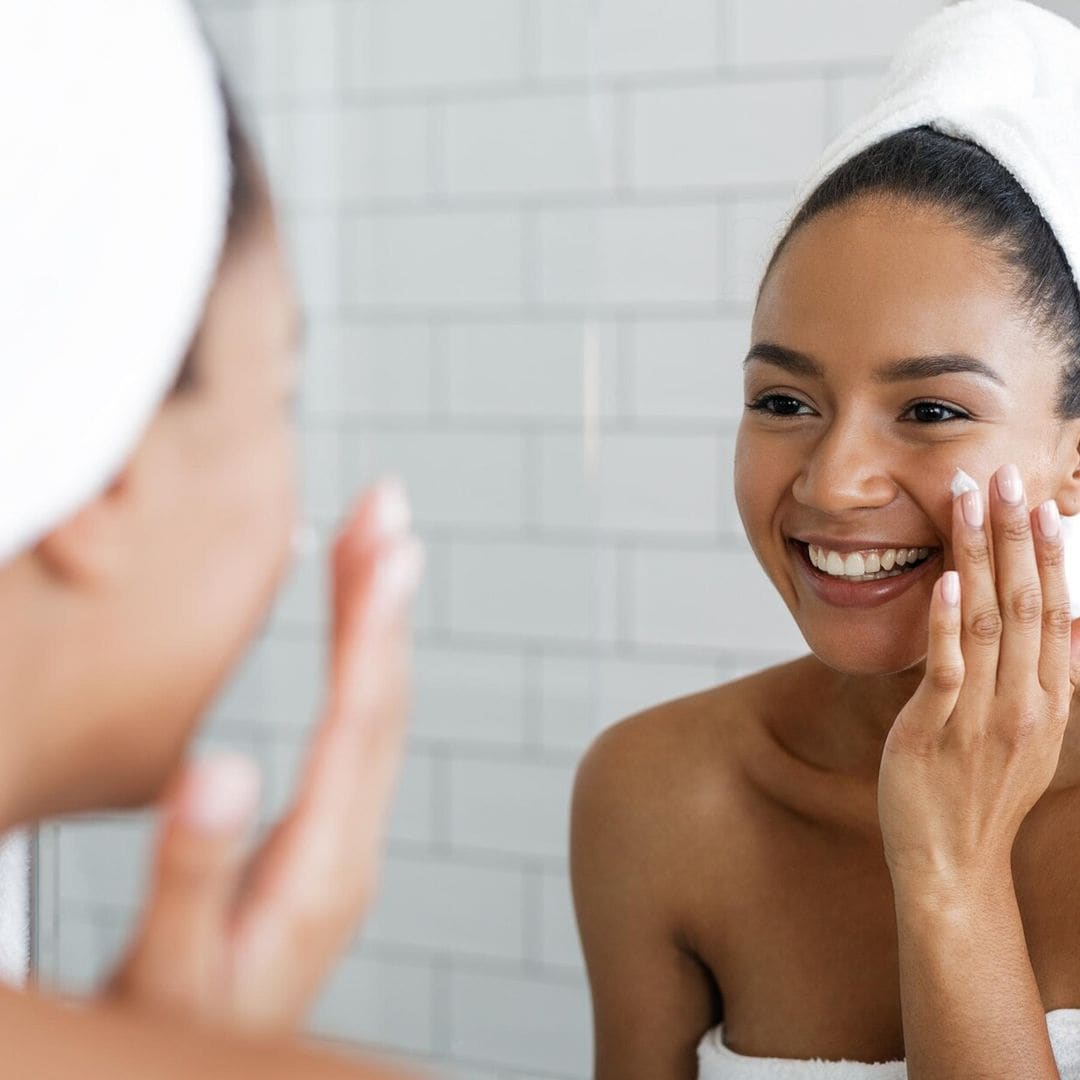 happy woman putting on facial moisturizer with hand in bathroom mirror