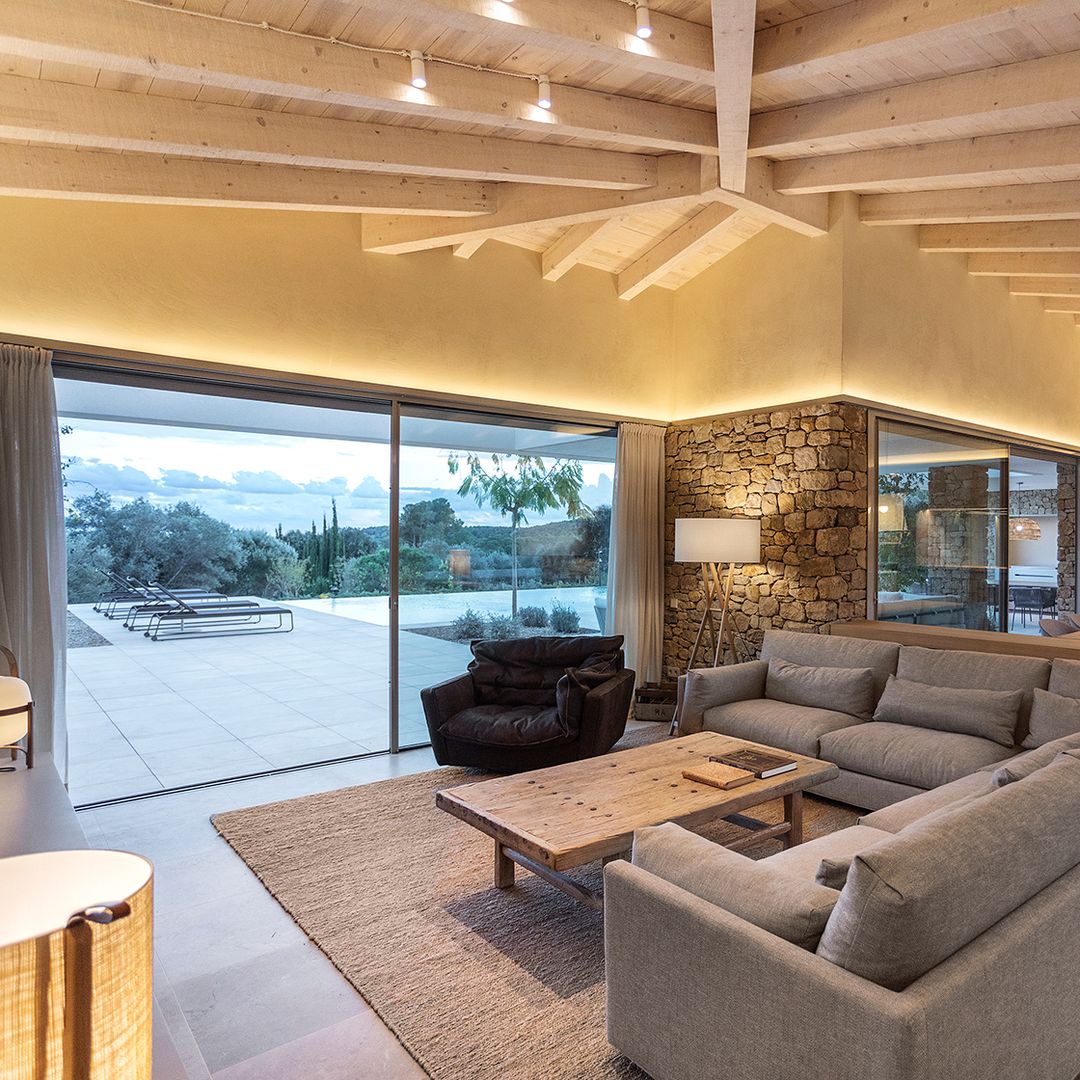 Salón comedor con pared de piedra, sofá gris y ventanal con vistas a la piscina 