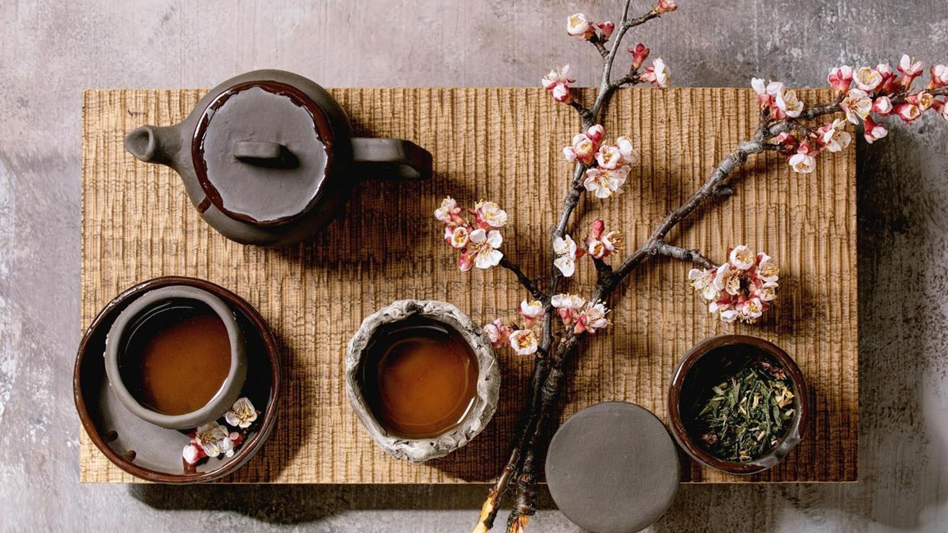 Tea drinking wabi sabi japanese style dark clay cups and teapot on wooden tea table with blooming cherry branches. Grey texture concrete background. Flat lay. space