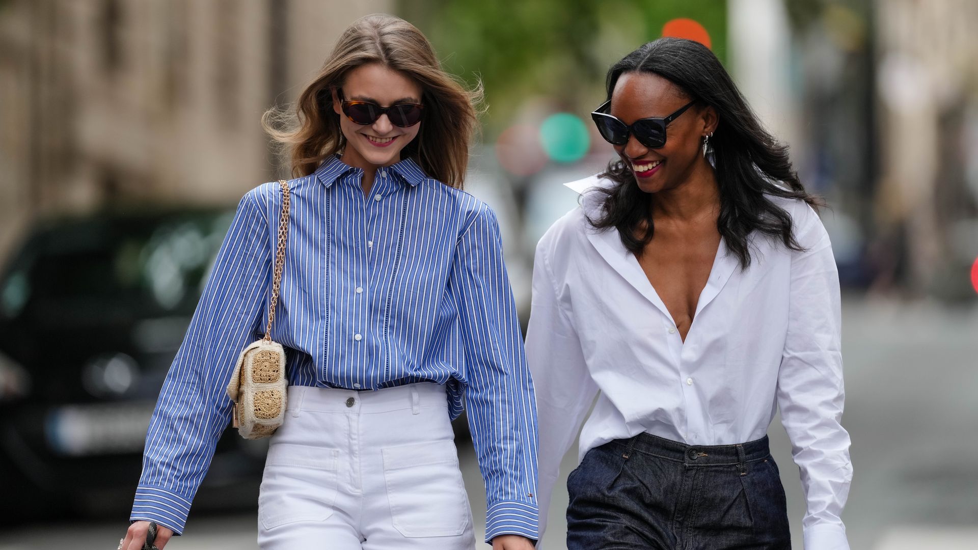 Segolene Hyppolite (L) wears sunglasses, a blue and white oversized shirt from Tommy Hilfiger, a raffia beige bag from Vanessa Bruno, high waist white denim pants from Vanessa Bruno; Emilie Joseph (R) wears a summer wardrobe capsule: an oversized white poplin button-down shirt from Figaret Paris, tucked into high-waisted denim bermuda/jorts/denim shorts, classic/basic/timeless accessories such as black sunglasses from the Korean fashion brand Gentle Monster, a small black top handle leather handbag,  during a street style fashion photo session, on May 20, 2024, in Paris, France. 