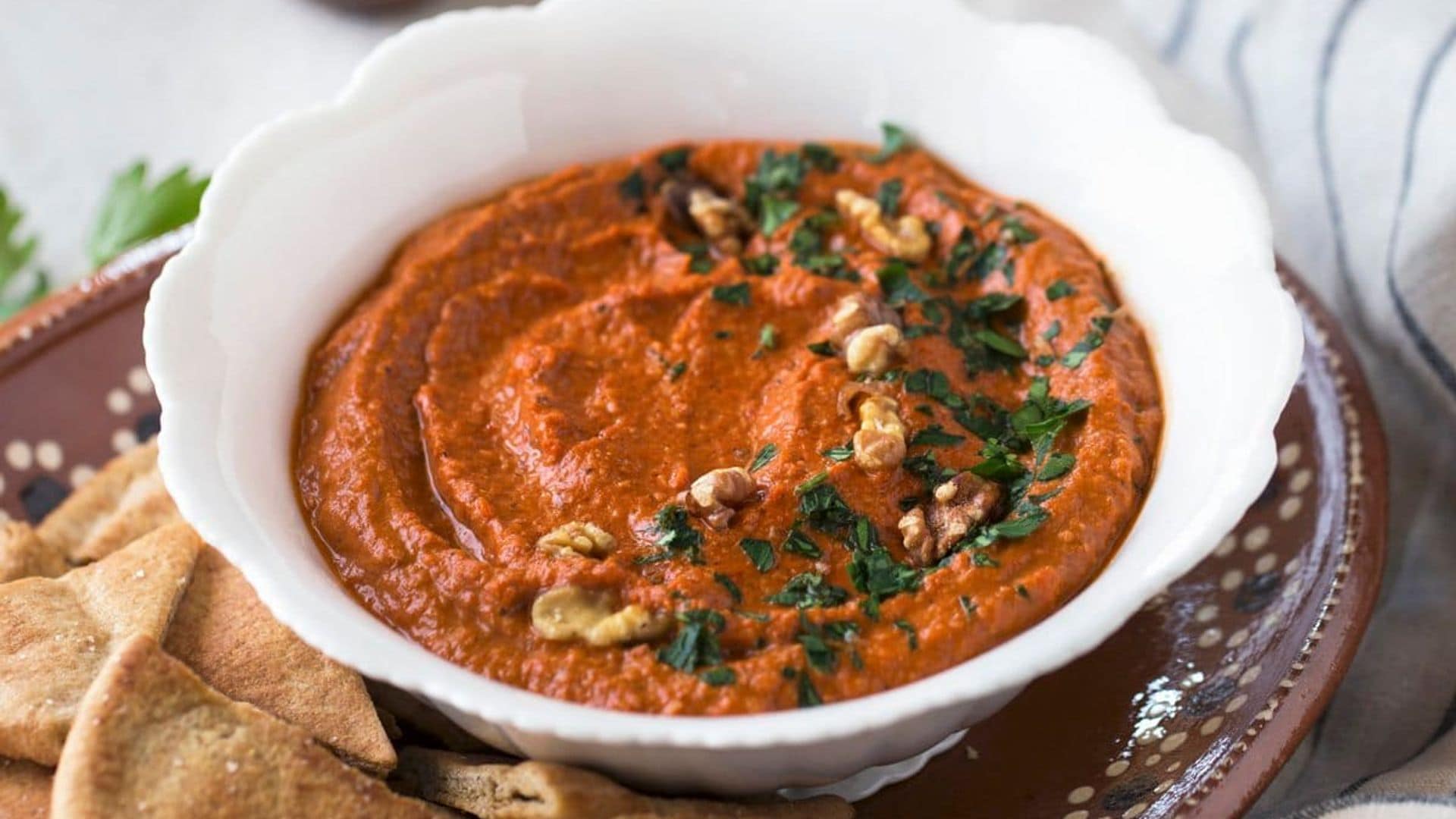 A bowl of red pepper and walnut dip served with pita chips on a plate