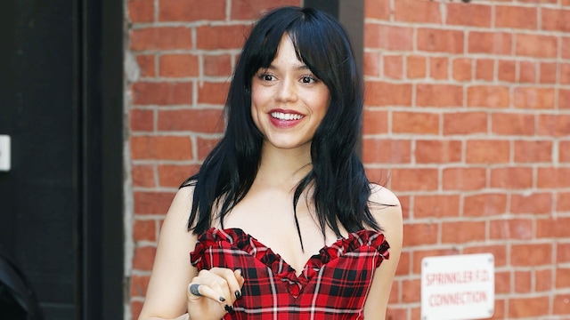 enna Ortega flashes a smile and waves as she steps out in New York City. The 21-year-old American actress strikes a fashionable pose in a red and black plaid dress paired with bright red heels.