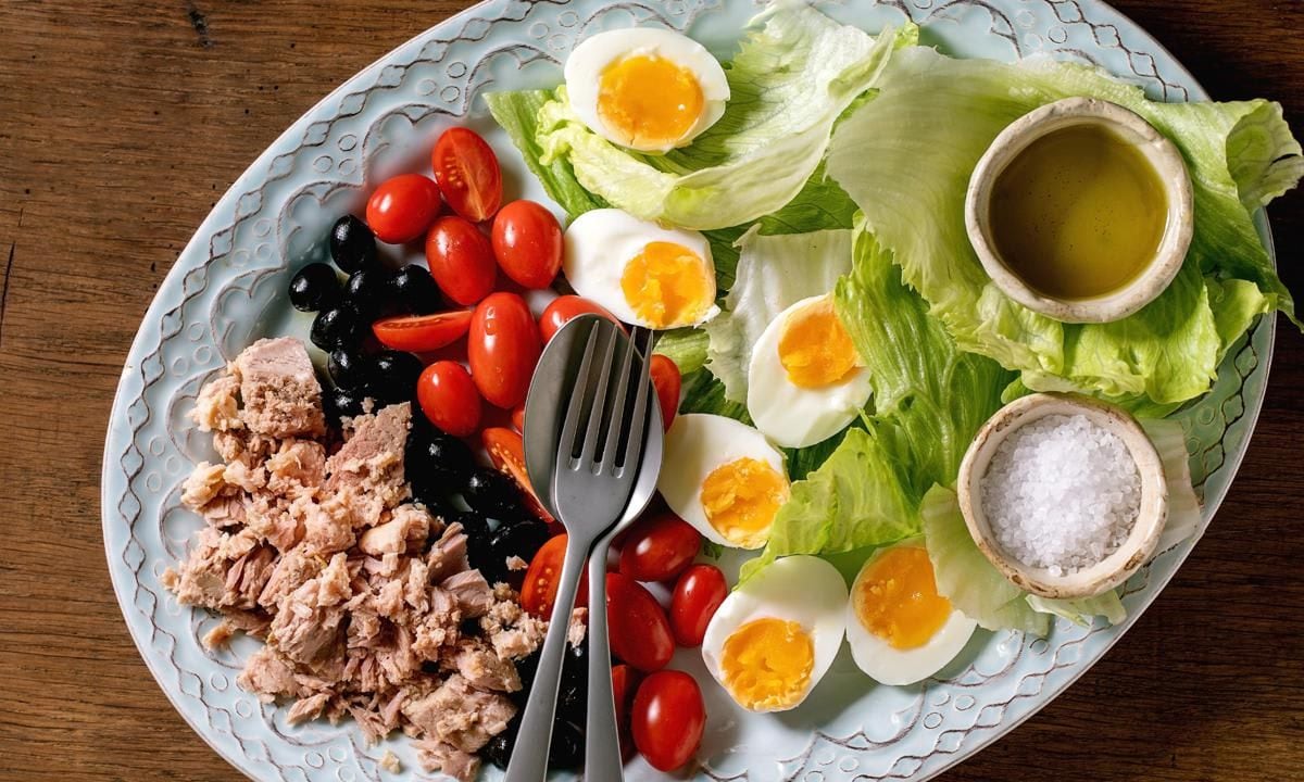 Layered french nicoise salad with canned tuna fish, olives and eggs, served with olive oil dressing and sea salt on ceramic plate over wooden background, Flat lay, copy space