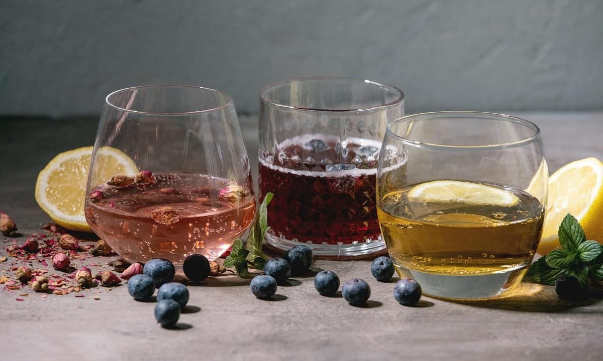 Different cocktail glasses with sparkling colorful drinks pink rose champagne, cider or lemonade with dry rose buds, lemon, blueberries and mint. Ingredients above. Gray texture background.